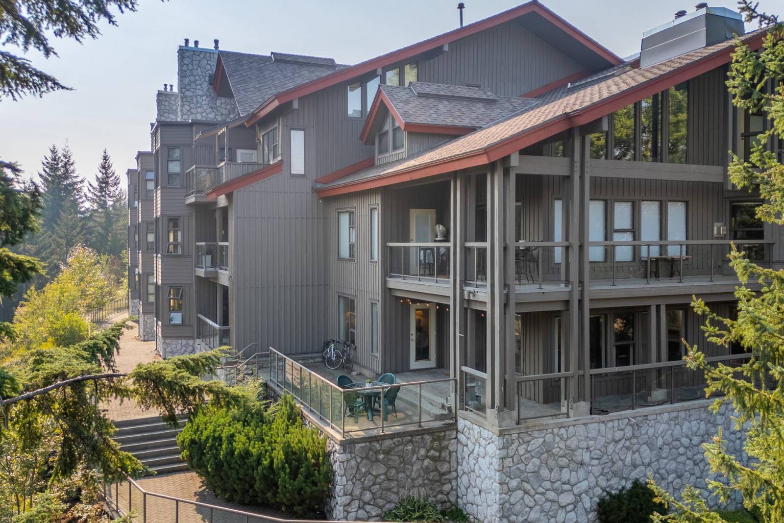 The Greyhawk building seen from the garden side, showing the ground-floor corner unit with its decks and stone base, framed by evergreens.