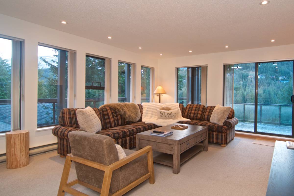 The living room with a deep sectional sofa and an armchair, wrapped by windows that frame the mountains, with patio doors to one side.